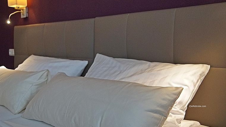 Une chambre moderne avec une tête de lit rembourrée et deux grands coussins blancs. Le mur est d'un violet foncé et un applique murale fournit une lumière douce.