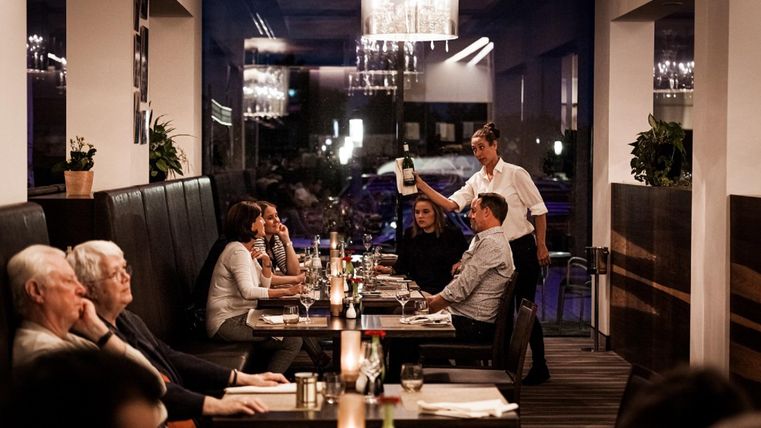 An elegant restaurant with a cozy atmosphere. Guests enjoy their meal while a waiter offers a wine.