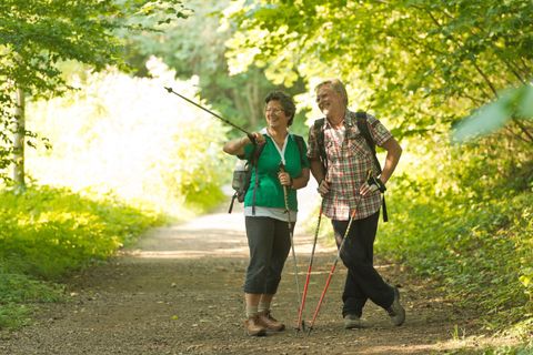 Ein glückliches älteres Paar wandert auf einem schmalen Weg im Wald. Sie tragen Rucksäcke und Wanderstöcke und genießen gemeinsam die Natur.