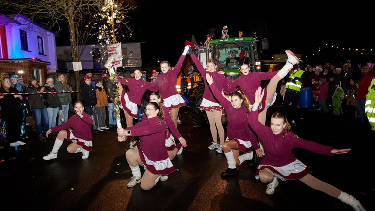 Een groep danseressen voert een act op in paarse kostuums onder de lichten van een nachtparade. Toeschouwers kijken enthousiast naar het spektakel.