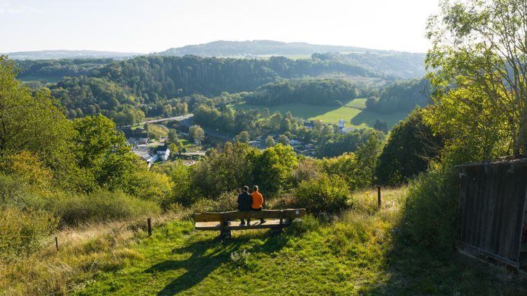 Een blik op een schilderachtig landschap met zachte heuvels en veel groen. Twee personen zitten op een bank en genieten van het uitzicht.