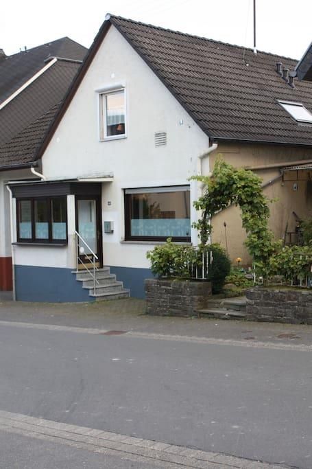 Ein gemütliches Einfamilienhaus mit einer kleinen Treppe und einem begrünten Vorgarten. Die Fassade ist in hellen Farben gehalten und es gibt Fenster mit Rollläden.