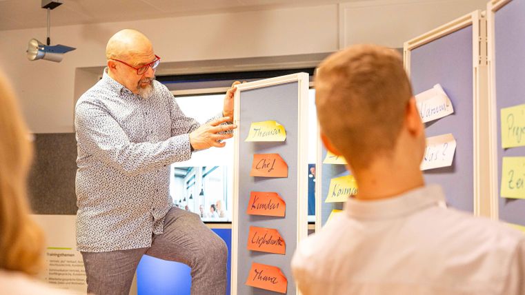 Ein Mann erklärt etwas an einem Präsentationsboard mit bunten Zetteln. Im Hintergrund ist eine Person zu sehen, die aufmerksam zuhört.