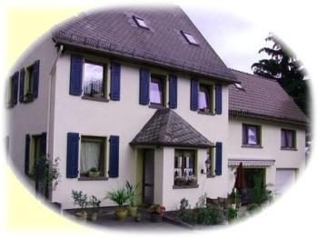 Une belle maison à deux étages avec un toit pointu et des volets bleus. L'entrée est accueillante, décorée de plantes.