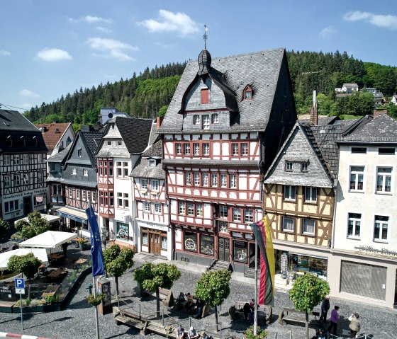 Historische Fachwerkh&auml;user am Marktplatz in Adenau, umgeben von B&auml;umen und Flaggen. Menschen sitzen und spazieren bei sonnigem Wetter., &copy; Stadt Adenau