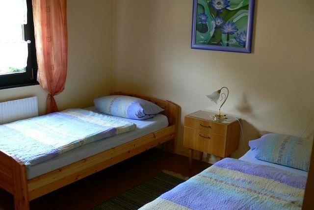 Een eenvoudige slaapkamer met twee eenpersoonsbedden en een nachtkastje. Aan de muur hangt een schilderij met blauwe bloemen, en het raam heeft oranje gordijnen.