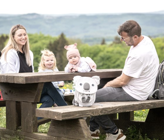 Eine Familie sitzt an einem Holztisch im Freien. Zwei Kinder, ein Kinderwagen und ein Rucksack sind zu sehen. Im Hintergrund ist eine grüne Landschaft., © TI Hocheifel-Nürburgring,D.Ketz