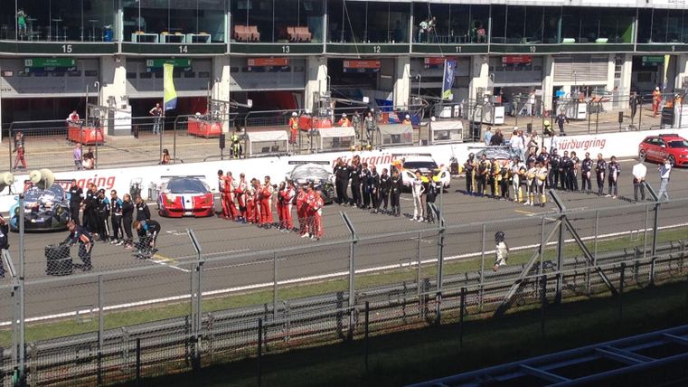 Een groep coureurs en teamleden verzamelt zich op het circuit. Op de achtergrond zijn de startopstellingen en de tribunes van het publiek te zien.
