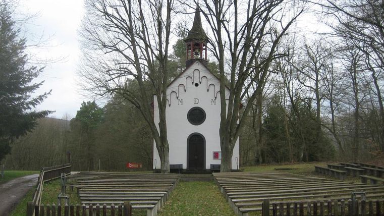 A small church surrounded by trees and a circular meadow. In front of the church, there are wooden benches for visitors.