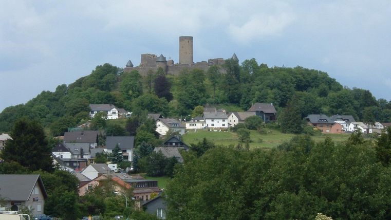 Un château sur une colline surplombe un petit village avec de nombreuses maisons. Le ciel est nuageux, et les environs sont verts et vallonnés.