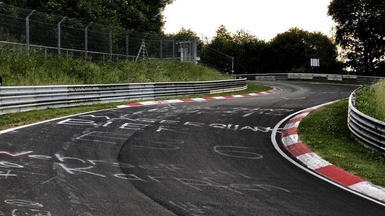 Eine geschwungene Rennstrecke mit Graffiti auf dem Asphalt. Im Hintergrund sind Bäume und ein Zaun sichtbar.