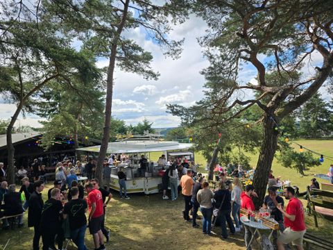 A lively outdoor event with many people gathered around a food truck. In the background, there are trees and a beautiful sky.