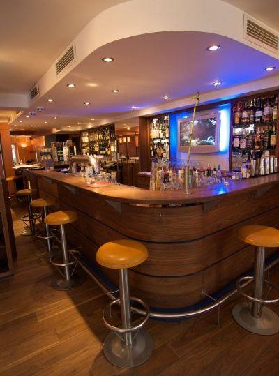 A stylish bar with a curved wooden counter and orange stools. In the background, there are shelves with various drinks and atmospheric lighting.