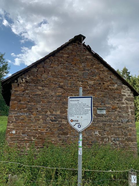 Ein altes Steinhaus mit einem begrünten Umfeld. Davor steht ein Schild mit Informationen über das Gebäude.