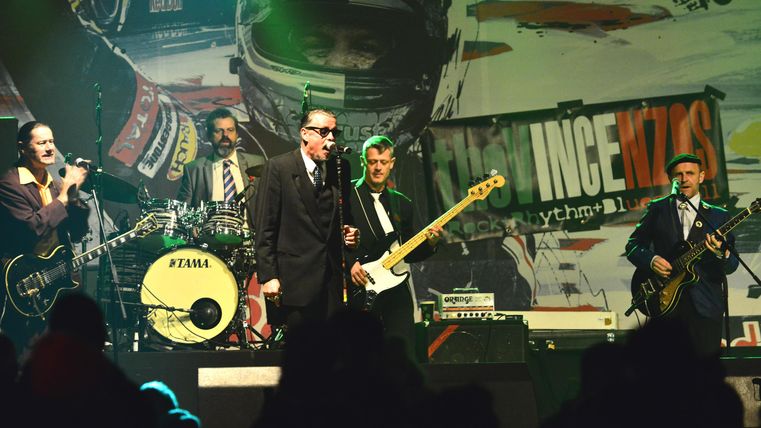 A band is performing live on stage against a colorful background. The musicians are wearing suits and playing various instruments.