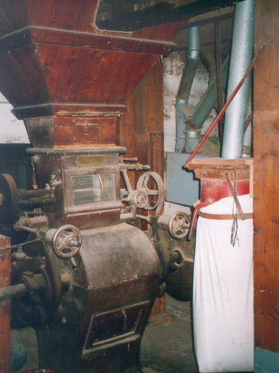 Eine alte Mahlmaschine in einem rustikalen Raum. Im Hintergrund sind Holz- und Metallstrukturen sichtbar.