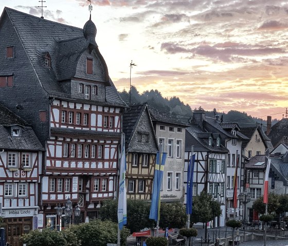 Fachwerkh&auml;user am Marktplatz in Adenau, beleuchtet von einem farbenfrohen Sonnenuntergang. Fahnen und B&auml;ume s&auml;umen die Stra&szlig;e., &copy; TI Hocheifel-N&uuml;rburgring, S.Schulte