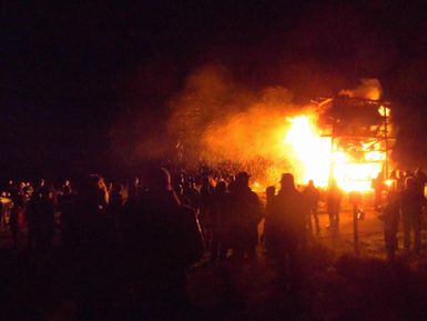 Eine große Menschenmenge steht vor einem lodernden Feuer, das ein Gebäude in der Dunkelheit beleuchtet. Der Himmel ist rauchig, und die Szenerie vermittelt ein Gefühl der Dramatik.