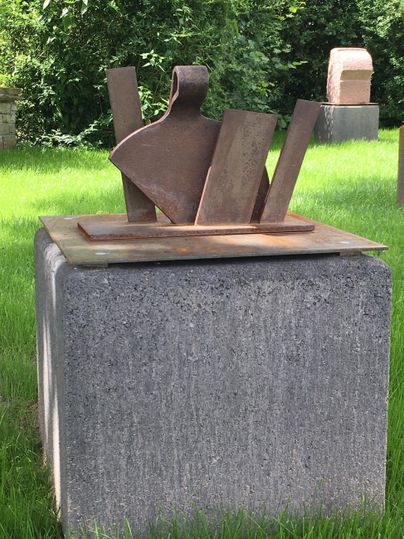 Une sculpture moderne en métal se dresse sur un bloc de pierre grise. Entourée d'herbe verte et d'arbres, elle semble contemporaine et attrayante.