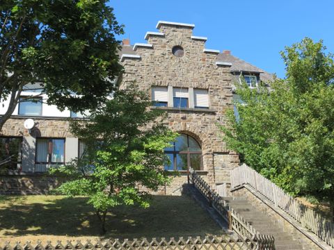Ein historisches Gebäude aus Stein mit großen Fenstern und einer markanten Fassade. Umgeben von Bäumen und einem kleinen Garten.