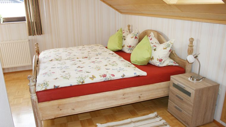 Ein gemütliches Schlafzimmer mit einem Holzbett und bunten Kissen. Der Boden ist aus Parkett und ein kleiner Nachttisch steht neben dem Bett.