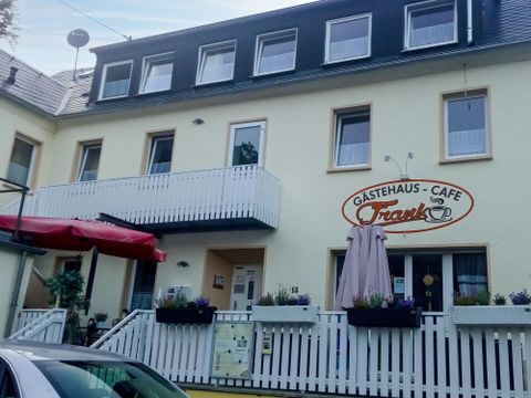 A cozy guesthouse with a café, decorated with a white fence wall and blooming plants. The entrance area is designed to be welcoming with a sunshade and outdoor tables.