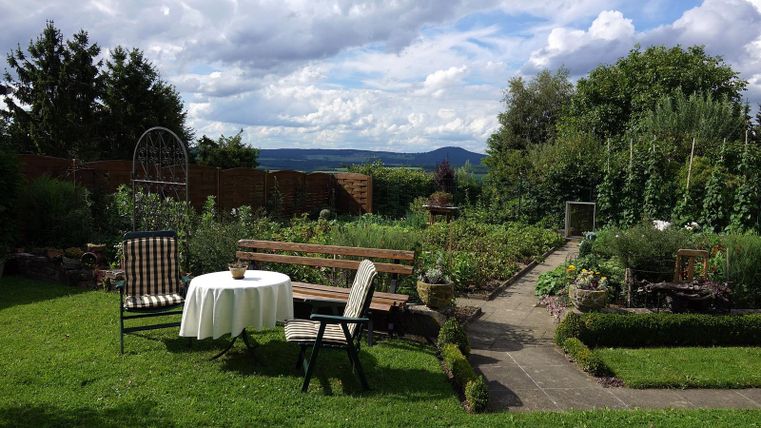 A beautiful garden with a table and chairs. In the background are plants and trees under a cloudy sky.