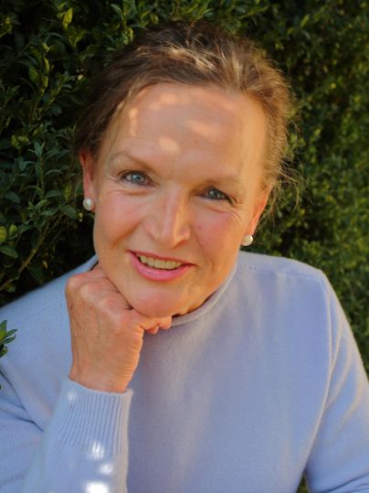 A woman with a smiling face is sitting in front of a green hedge. She is wearing a light blue sweater and has one hand resting on her chin.