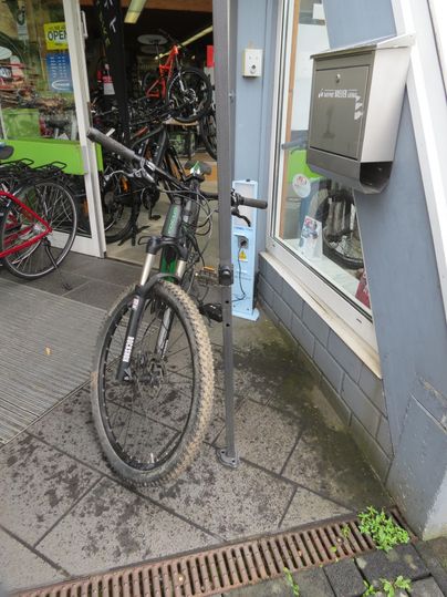 Ein Fahrrad steht an einem Fahrradständer vor einem Geschäft. Im Hintergrund sind weitere Fahrräder sichtbar.