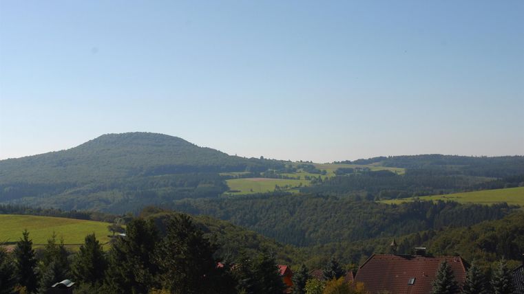 Een uitgestrekt, groen landschap met zachte heuvels en een heldere lucht. Op de voorgrond zijn enkele bomen en daken te zien.