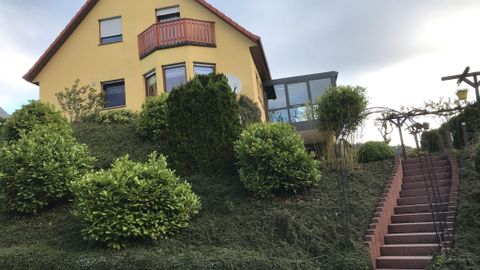 Une maison jaune se dresse sur une colline, entourée de buissons verts. Un escalier descend la colline.