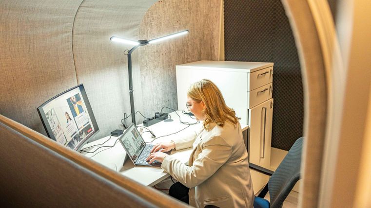 Eine Person arbeitet an einem Laptop in einem modernen Arbeitsraum. Der Raum ist klein und gut beleuchtet, mit einem Schreibtisch und einem Aktenschrank.