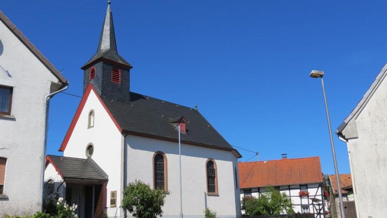 Eine kleine Kirche mit einem markanten Dach und einem steilen Kirchturm. Die Umgebung ist friedlich mit Wohnhäusern und einem klaren blauen Himmel.