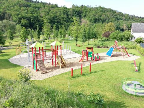 Ein Spielplatz mit bunten Geräten, darunter Rutschen und Schaukeln, in einer grünen Umgebung. Im Hintergrund sind Bäume und eine sanfte Hügelkette zu sehen.
