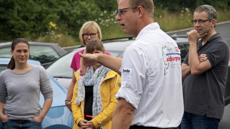 Eine Gruppe von Menschen hört einem redenden Mann in einem weißen Hemd zu. Im Hintergrund sind Autos und eine grüne Landschaft sichtbar.
