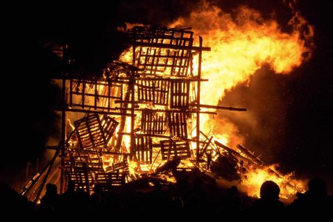 Ein großes Feuer brennt in einer zerstörten Holzkonstruktion. Menschen stehen in der Nähe und beobachten das Inferno.