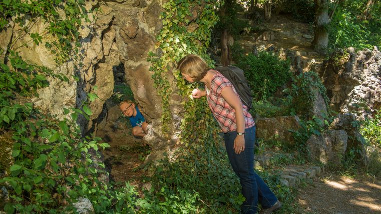 Eine Frau mit Rucksack schaut in eine mit Efeu bewachsene Felsöffnung, in der ein Kind sitzt.