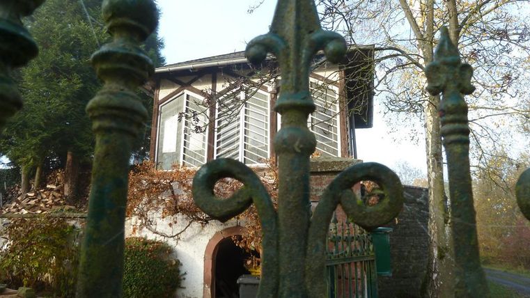 An old, weathered building behind an iron gate. Surrounded by trees and overgrown grass.