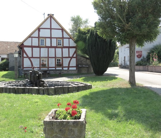 Fachwerkhaus mit rotem Holzrahmen, Brunnen und Blumenbeet im Vordergrund, Baum und Stra&szlig;e rechts, sonniger Tag., &copy; TI Hocheifel N&uuml;rburgring