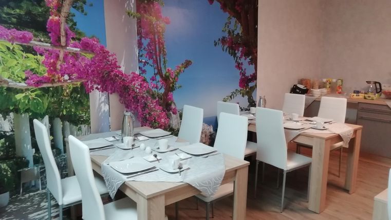 An inviting dining area with white chairs and wooden tables. The wall is decorated with a beautiful mural of colorful flowers and a landscape.