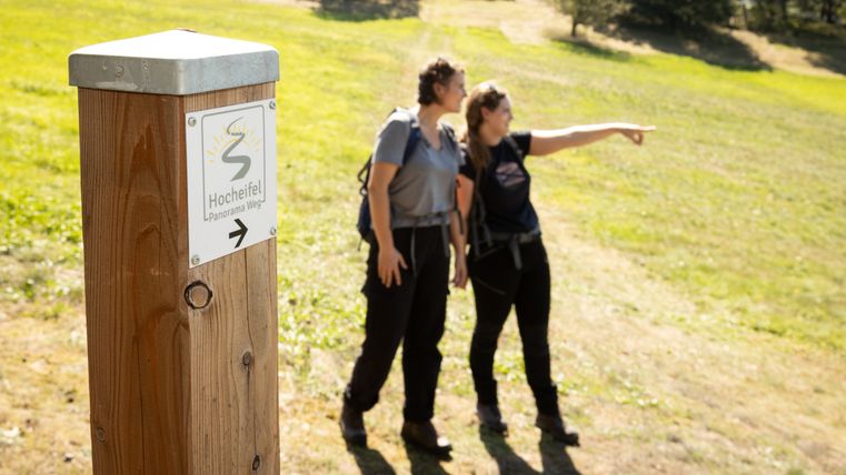 Zwei Personen auf einem Wanderweg in der Hocheifel, eine zeigt in die Ferne. Ein Wegweiser mit der Aufschrift 'Hocheifel Panorama Weg'.