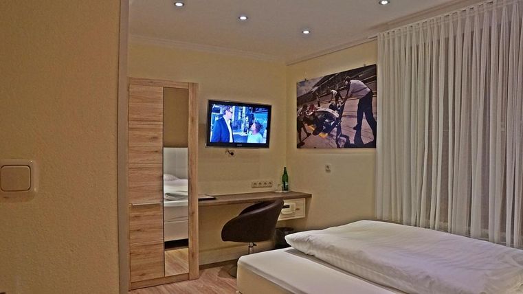 Une chambre d'hôtel moderne avec un lit, un bureau et une télévision. Les murs sont peints en clair et il y a de grandes fenêtres avec des rideaux.