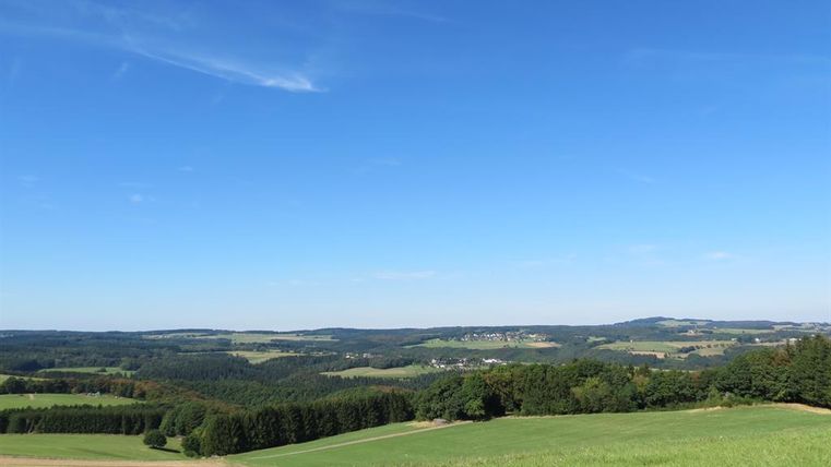Eine weite, grüne Landschaft unter einem klaren blauen Himmel. Sanfte Hügel und Wälder erstrecken sich bis zum Horizont.