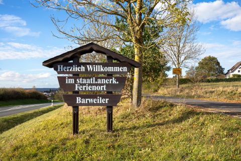 Ein Willkommensschild steht an der Straßenseite. Im Hintergrund sind sanfte Hügel und ein blauer Himmel zu sehen.