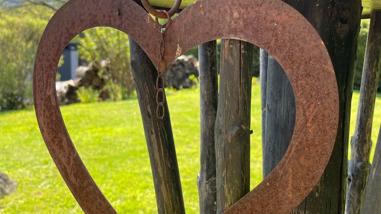 Un objet en métal rouillé en forme de cœur pend à un poteau en bois. En arrière-plan, on voit de l'herbe verte et des arbres.