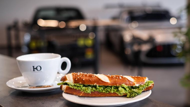 Ein frischer Salat-Sandwich auf einem Teller neben einer Tasse Kaffee. Im Hintergrund sind klassische Autos sichtbar.