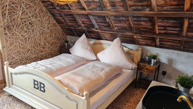 Een gezellige slaapkamer met een tweepersoonsbed en een rustieke houtdesign. De muren zijn gedeeltelijk bekleed met stro, en er is een kleine wastafel naast het bed.