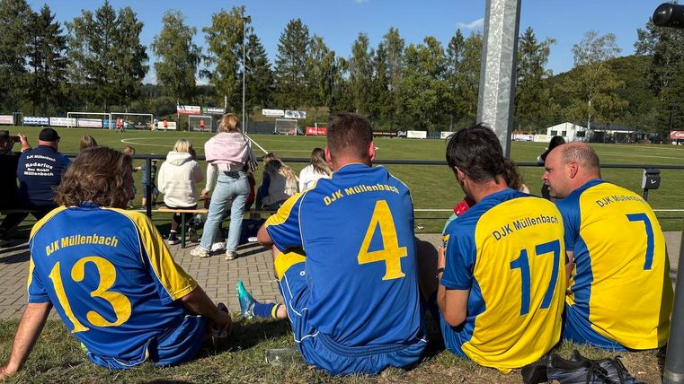 Eine Gruppe von Fußballspielern sitzt auf dem Boden und schaut auf das Spielfeld. Die Spieler tragen blaue und gelbe Trikots des DJK Mittenbach.