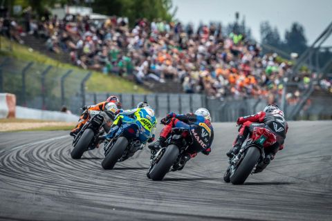 Eine actionreiche Motorradrennveranstaltung auf einer Rennstrecke. Mehrere Fahrer fahren in enger Formation, während begeisterte Zuschauer im Hintergrund zuschauen.