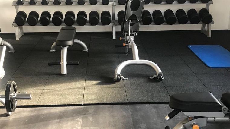 A fitness room with a barbell and several dumbbells on a shelf. The floors are covered with mats, and there are a few fitness machines.